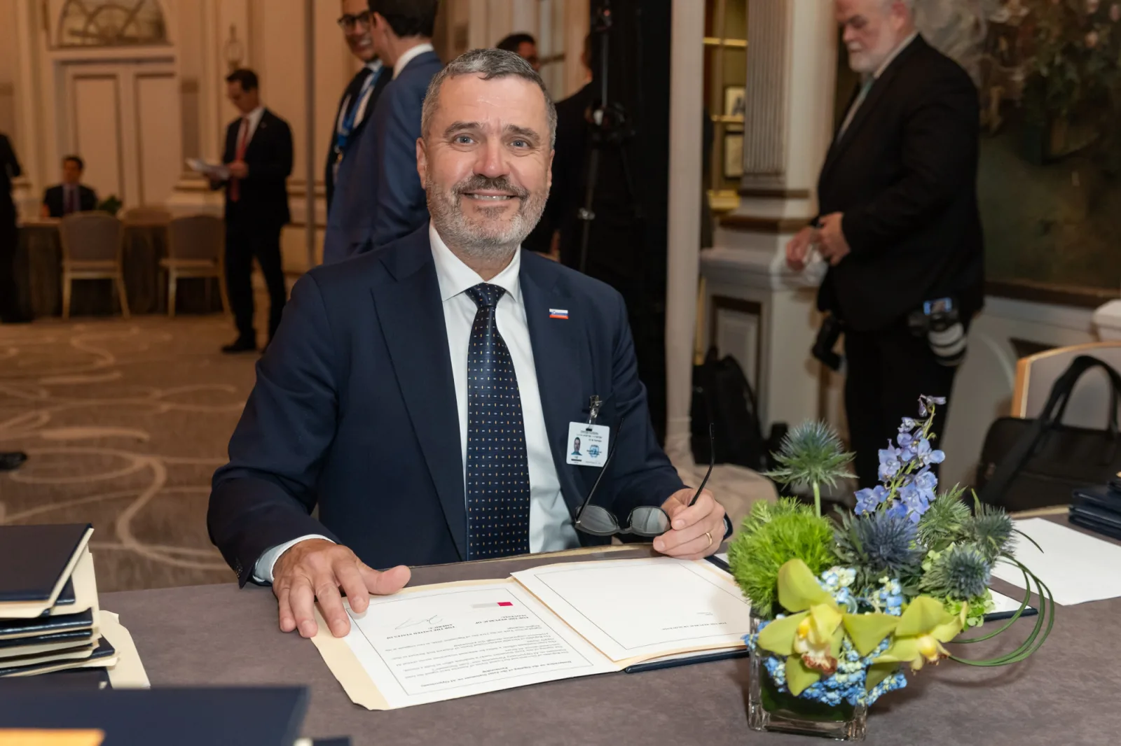 A suited man wearing a Slovenian flag pin signs a document at a formal event, with a flower arrangement in the foreground.