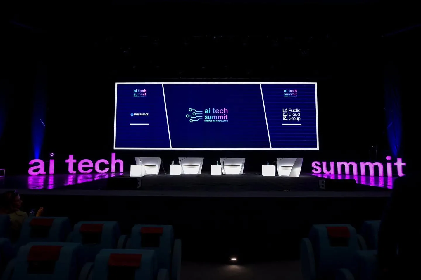 Empty AI Tech Summit stage with four panel chairs, a branded screen and magenta-lit lettering.