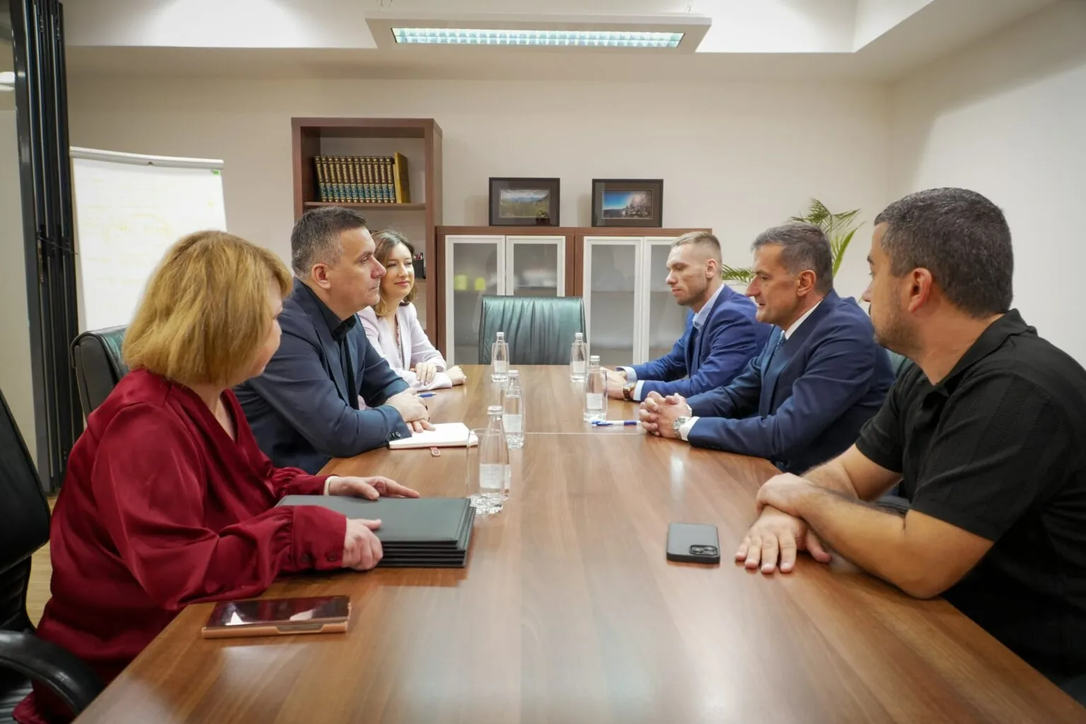 Six people in business attire meet around a conference table with water bottles and phones.